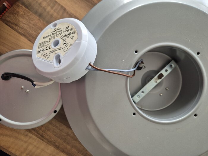 Elektronisk transformator ansluten till en grå taklampa med synliga kablar och kopplingsbox på ett träbord.