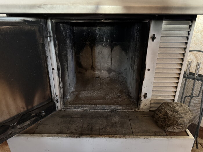 Öppen eldstad med sotiga väggar; dörren till eldstaden är lite öppen och en stor sten ligger på spisen.
