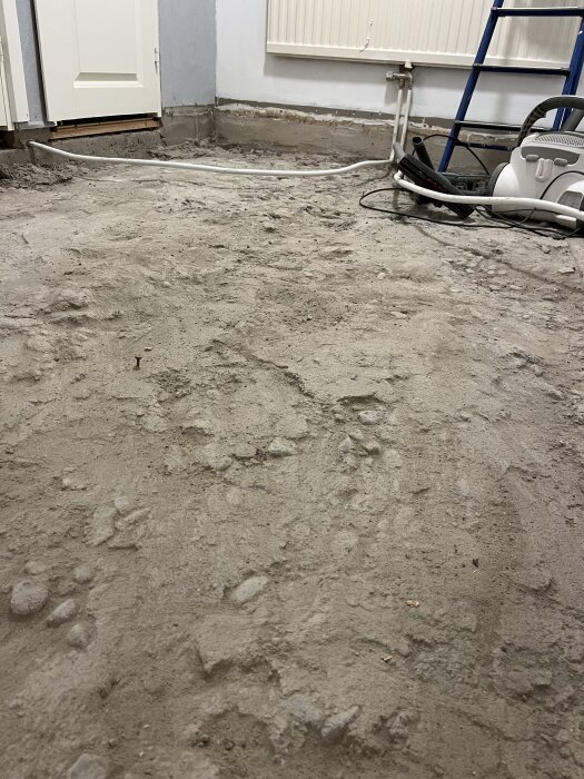 Betonggolv med ytskikt av sand och damm i ett rum under renovering, med en stege och dammsugare i bakgrunden.