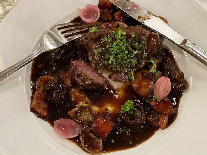 Tallrik med beuf bourgogne, garnerad med persilja och syltlök, serverad med kniv och gaffel på en vit tallrik.
