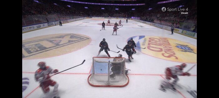 Ishockeymatch med spelare i rödvita och mörka dräkter på isen, puck nära målområdet och åskådare i bakgrunden.