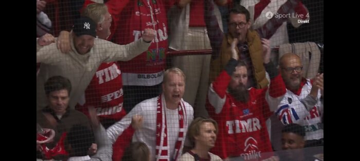 Hockeyfans jublar bakom ett skyddsnät, klädda i röda och vita Timrå-tröjor. De ser glada och uppspelta ut under ett sportevenemang.