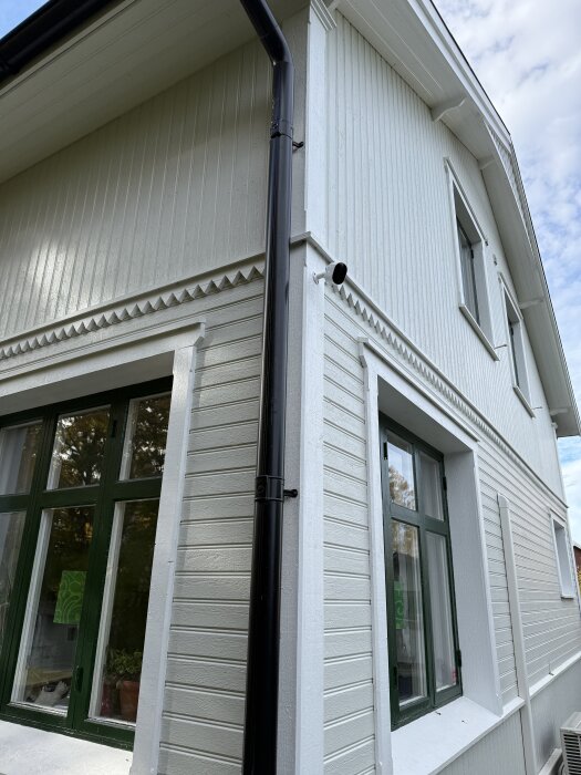Vit husvägg med monterad kamera nära taket, bredvid ett svart stuprör. Grönt fönster syns på nedervåningen.