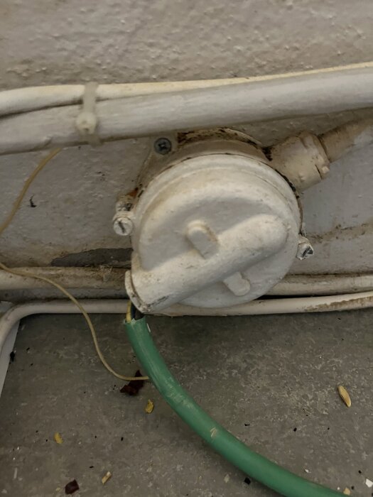 Väggmonterad vit anslutning för spis med grön elkabel och synliga skruvar, placerad i en äldre hyresrättsmiljö.