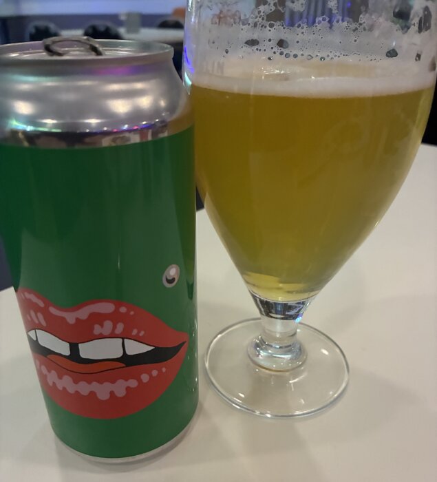 Grön dryckesburk med rött läppmotiv bredvid ett glas gyllene öl med skum, står på ett bord.
