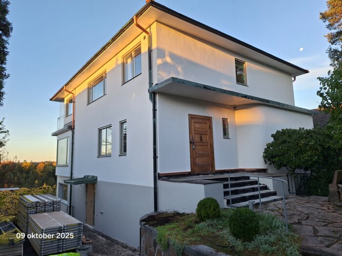 Nyrenoverad husfasad med vit puts, bruna dörrar och fönster, omgiven av grönska och byggmaterial i förgrunden, i kvällssol.