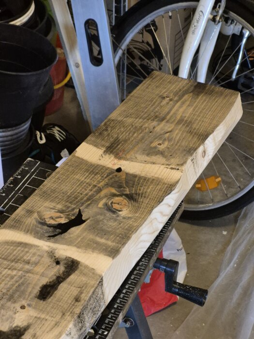 Bräda av furu med mörkbehandlad yta, klämd på ett arbetsbord i ett garage, omgivna av andra verktyg och cykeldäck.
