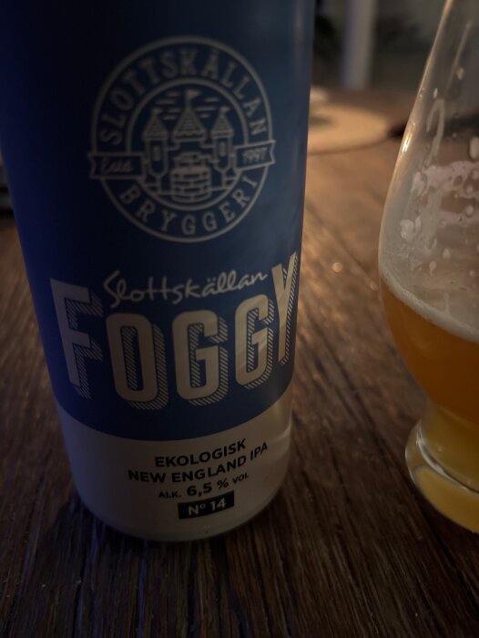 Blå burk med texten "Slottskällan Foggy, Ekologisk New England IPA, 6,5%". Glas bredvid med halvfull gul-orange vätska.