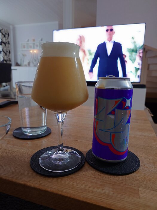 En öl från Omnipollo i ett glas bredvid en burk med färgglad etikett på ett bord i ett vardagsrum med en påslagen TV i bakgrunden.