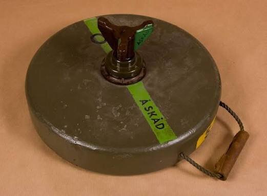 Gammal svensk mina med handtag och texten "ÅSKÅD" på en grön remsa över en brun bakgrund.