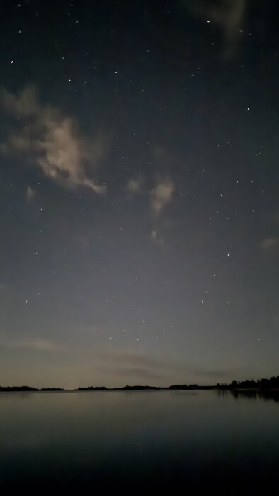 Stjärnklar natthimmel över en spegelblank sjö med mörka silhuetter av träd och horisont.