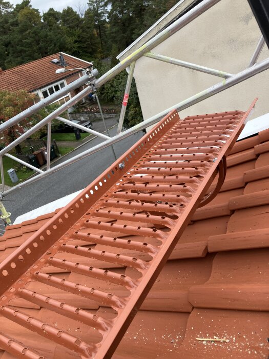 Takmonterad gångstege i metall över tegelpannorna, omgiven av byggställning och träd i bakgrunden.