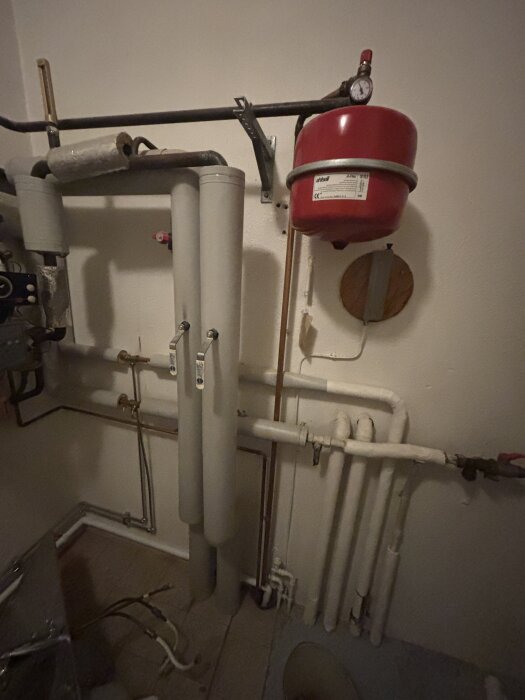 Pannrum med rör och röd trycktank på vägg. Tratt i golvet där vatten från fjärrvärmen droppar. Planerad anslutning för tvättmaskin.