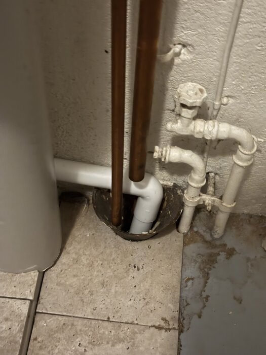 Tratt i golvet med kopparrör och plastavloppsrör i ett pannrum, potentiell anslutning för tvättmaskinsavlopp, omgiven av kakelgolv.