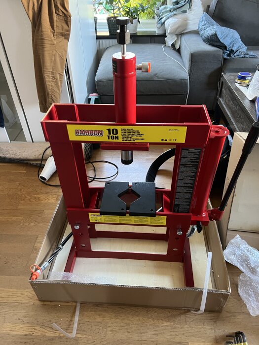 Röd hydraulpress med 10 tons kapacitet i ett vardagsrum, omgiven av verktyg och möbler.