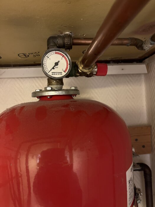 Manometer och röd trycktank med kopparrör i ett värmesystem. Manometern står på 0, och en röd ventil syns på höger sida av röret.