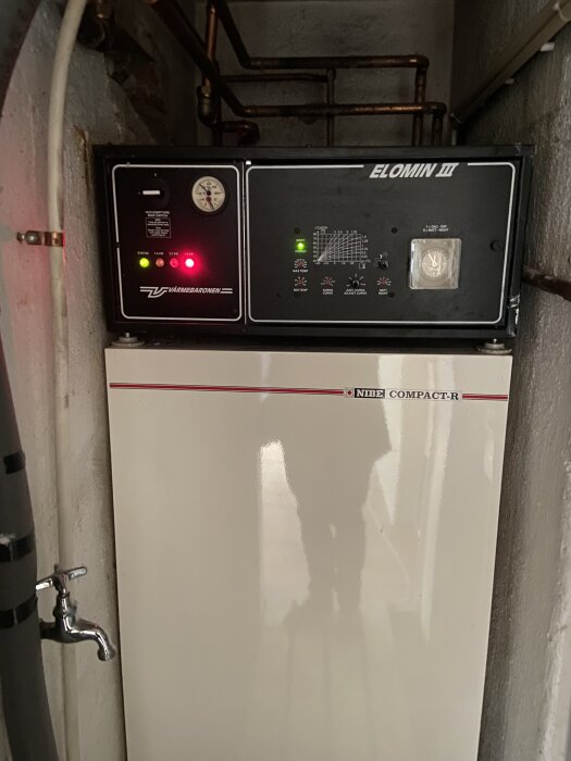 Värmepanna av märket Nibe Compact-R med tydliga kontrollpaneler och rörkopplingar, installerad i ett äldre hus med vattenburet värmesystem.