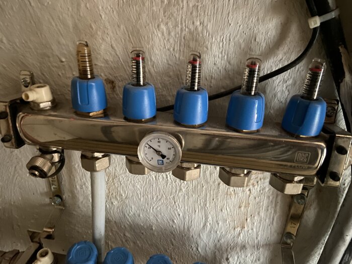 Värmesystemets fördelningsrör med fem blå ventiler och en tryckmätare monterad på en vägg, använd för ett vattenburet system.
