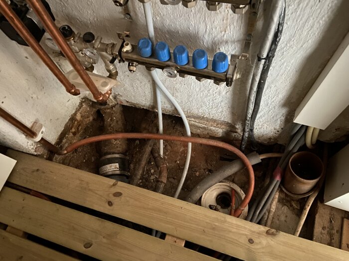 Vattenburet värmesystem med kopparrör och plastkopplingar synligt under en trätrall i ett äldre hus. Flera ledningar och rör synliga mot väggen.