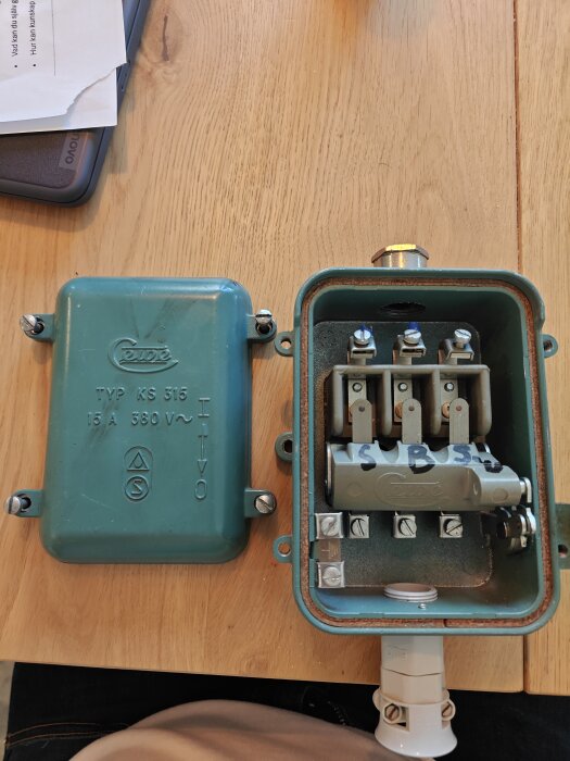 Öppen turkos kopplingsbox med synliga anslutningar och skruvar på träbord.