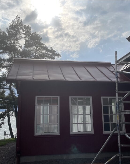 Ojämn bandplåtstak på ett nybyggt rött hus med ställning bredvid, solen bakom moln i bakgrunden.