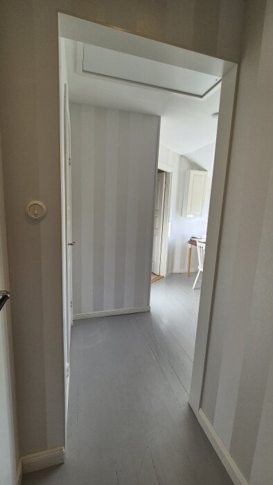 Hall med ljusgrått trägolv, randiga väggar och en dörröppning in till ett rum med ett bord och en stol.
