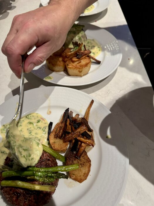En tallrik med potatisfondant, grillad entrecote, sparris och palsternacka strips. En hand skedar sås över rätten.