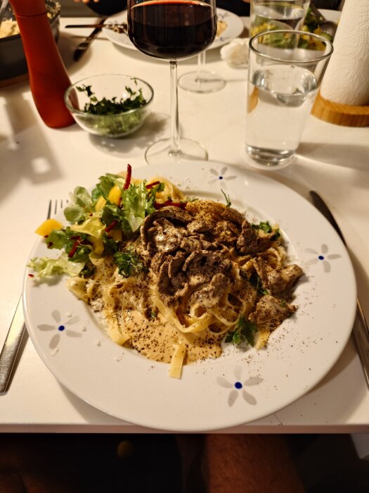 Tallrik med lövbiffssås och tagliatelle, toppad med parmesan och grönsaker, serverad med ett glas rött vin och vatten.
