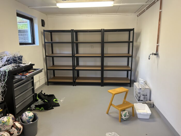 Hylla monterad i förråd, omgiven av verktygslåda, fotbollar och en gul pall; synliga vattenledningar.