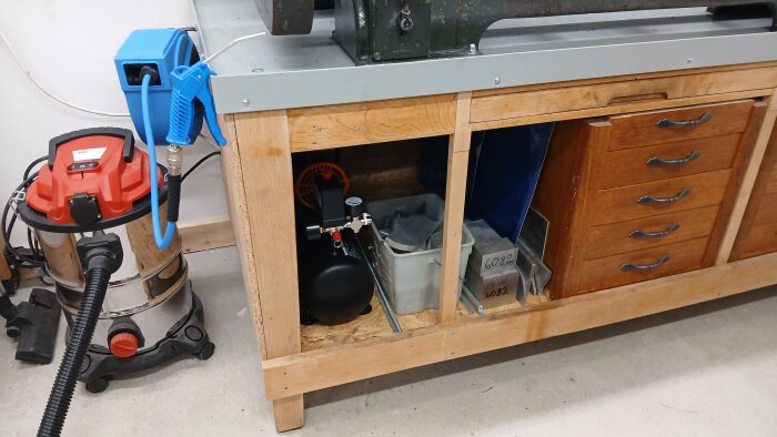 Tryckluftsutrustning och dammsugare i ett garage, placerad intill en arbetsbänk med förvaringslådor.