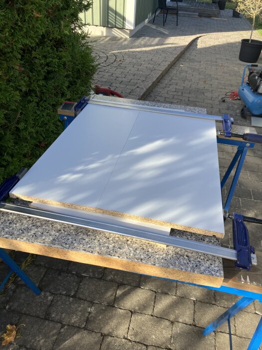 Två hyllplan i olika storlekar och färger på arbetsbänk utomhus, med verktyg och en kompressor i bakgrunden.