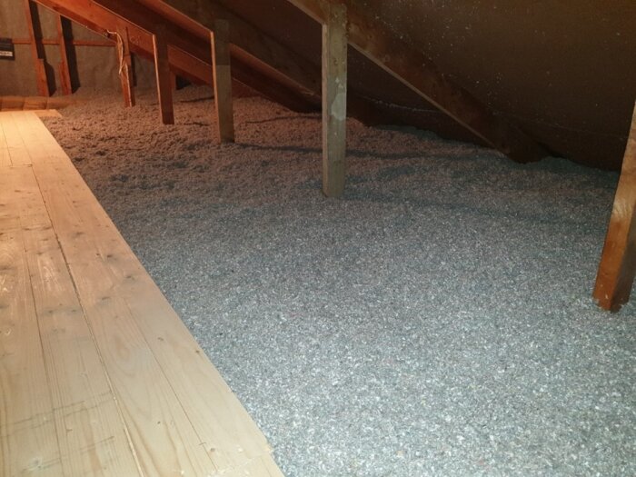 Vind med tjockt lager lösull och smal gång i mitten, brädor till vänster och träbalkar ovanför.