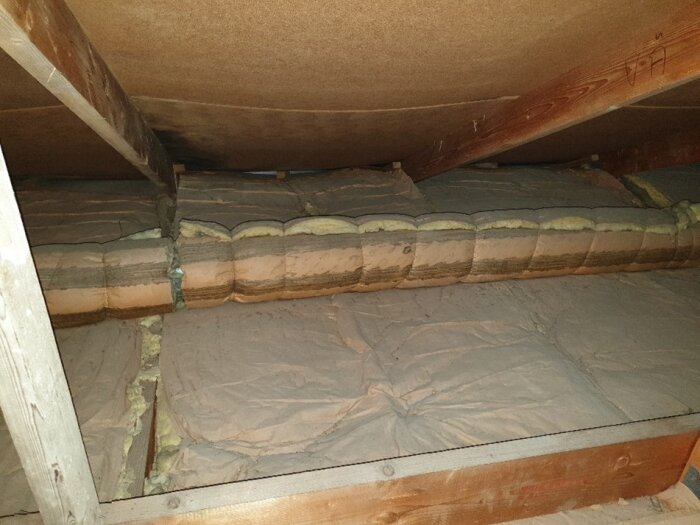 Bild av vinden med isoleringsmaterial av glasfiberull täckt med papp. Lösullsisolering ovanpå, träram synlig, snedtak ovanför.