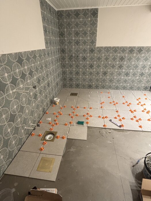 Halvlagt badrumsgolv med stora kakelplattor och karmar, väggarna har mönstrade kakelplattor. Planerade fogar syns mellan plattorna.