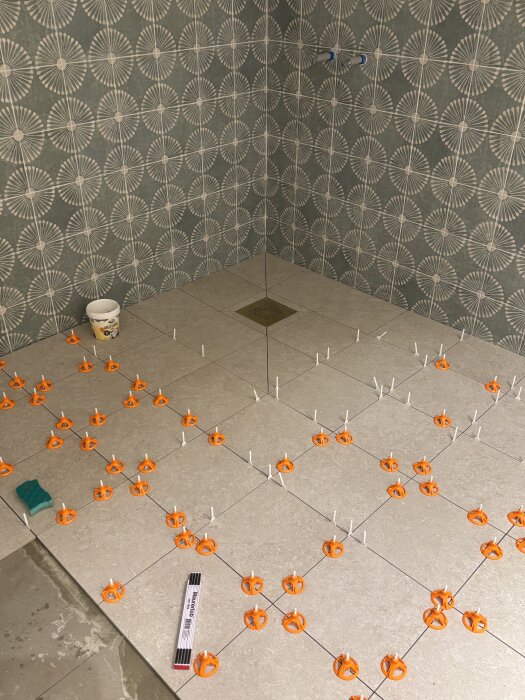 Klinkergolv under läggning i badrum, med orange kakelkryss och grönt väggkakel mönstrat med vita cirklar. Verktyg och hink på golvet.