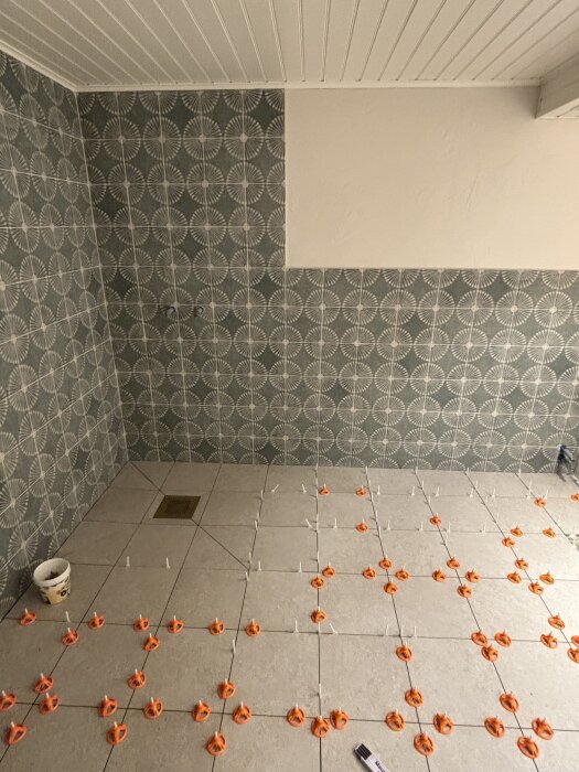 Golvläggning pågår i badrum, ungefär hälften färdigt. Plattor monteras med distanser, väggarna har grönt mönster. Tom målarhink synlig.