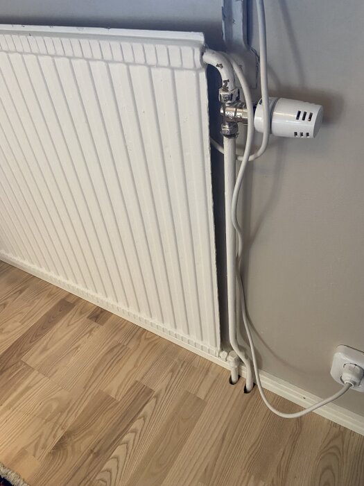 Radiator med smal design och anslutna rör på 18 mm. Placering mot trägolv och vägg med termostatventil synlig.