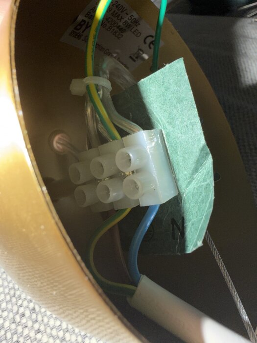 Elektrisk kopplingsdoskopplad i taket med synliga vita, blå och gulgröna kablar; grönt papper i takdosan.