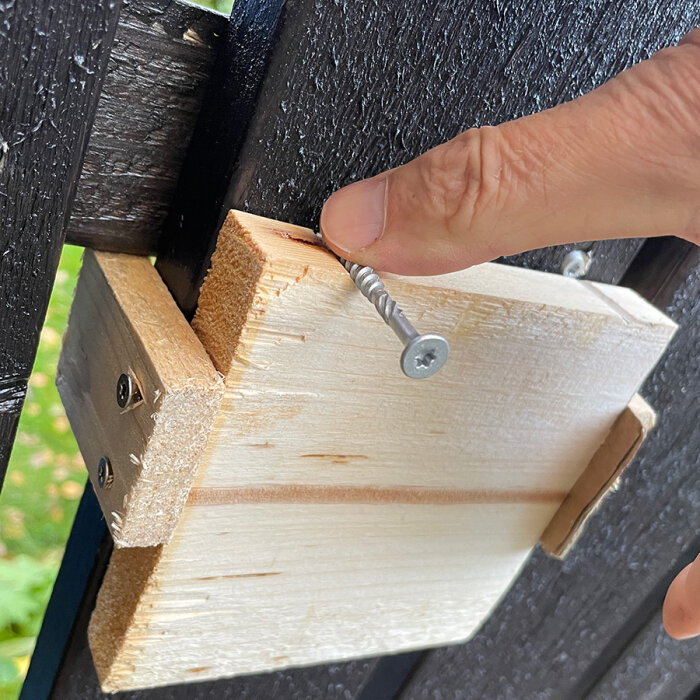 En hand håller en träbräda mot ett staket medan en skruv förs in genom en mall för att markera korrekt position.