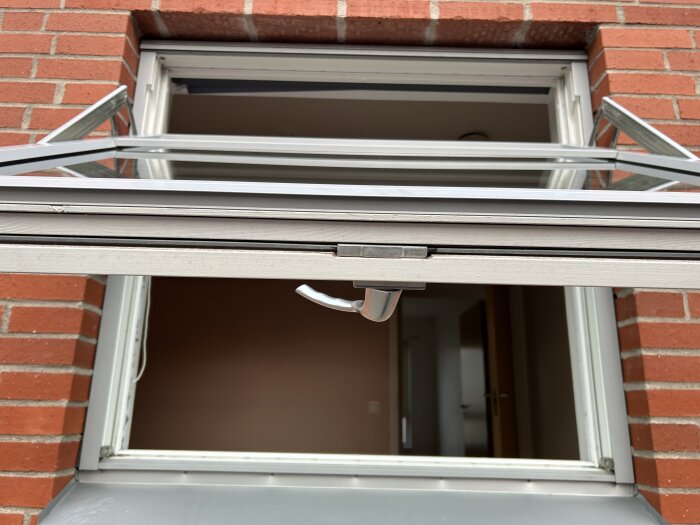 Fönster öppet utåt med synlig spanjolett och handtag, omgiven av röd tegelvägg.