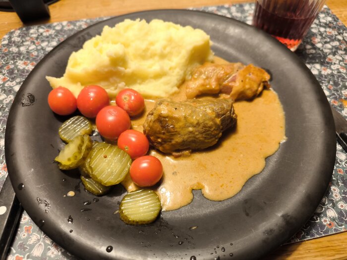 Baconlindade rullader med gräddsås, hemgjort potatismos, körsbärstomater och skivade pickles på en svart tallrik.