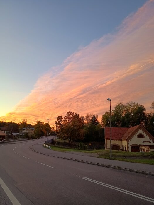 Utsikt över väg och kraftstation vid solnedgång i Skogstorp, med färgstarka moln och träd i bakgrunden.