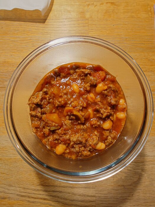 Skål med chili con carne med köttfärs, bönor och tomatbitar på ett träbord.
