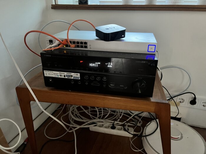 Förstärkare, nätverksutrustning och Apple TV på ett bord med kablar runtom, dammsugare synlig på golvet bredvid.