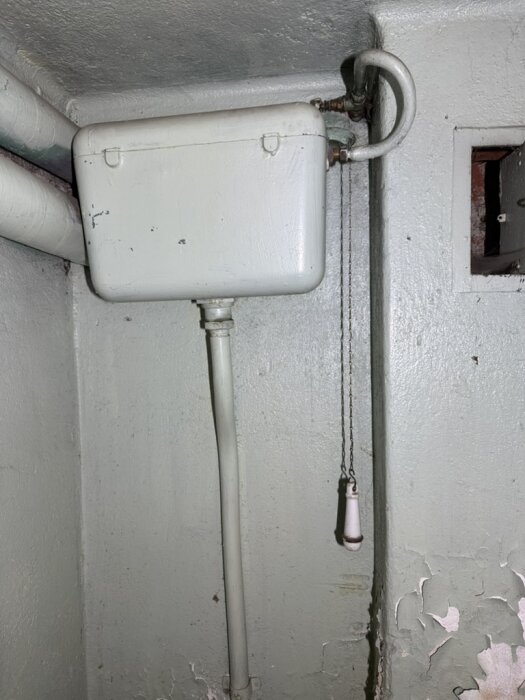 Gammal toalettsisterntank med kedja i ett hus från 1935, monterad högt upp på en vägg i en källare.