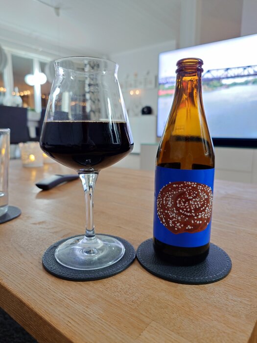 En ölflaska med en blå etikett och illustrerat bakverk bredvid ett glas öl på ett bord, med en TV i bakgrunden.