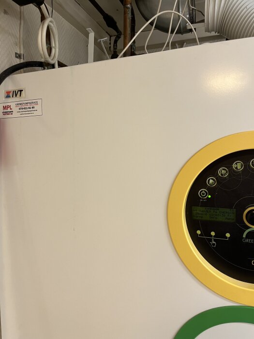 Panel på värmepump med display och om/on knappar, del av rörsystem och etikett med serviceinformation syns i bakgrunden.