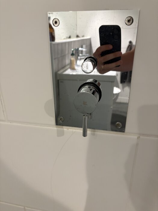 Duschblandare monterad på en vit kaklad vägg, med två vred och en spegelreflektion av en hand som håller en mobiltelefon.