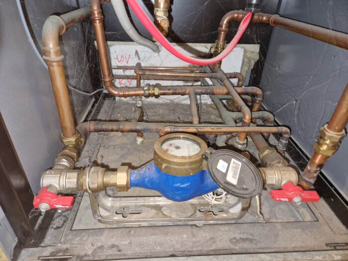 Vattenrör och ventilinstallation under diskbänk, med kopparrör och två röda ventiler. Spindelväv synlig i hörnen.