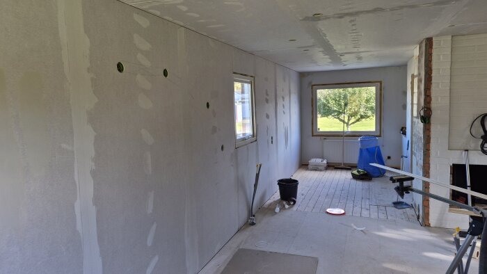 Vardagsrum under renovering med uppsatta och spacklade gipsskivor, fönster och verktyg synliga.